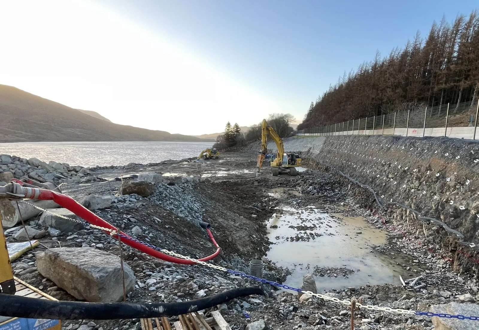 Excavators working in a deep, soil-nailed cutting beside a reservoir, with forest above